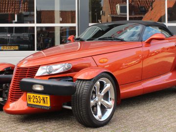 Een rode Plymouth Prowler-cabriolet met chromen wielen staat sierlijk geparkeerd voor een garage met grote ramen.