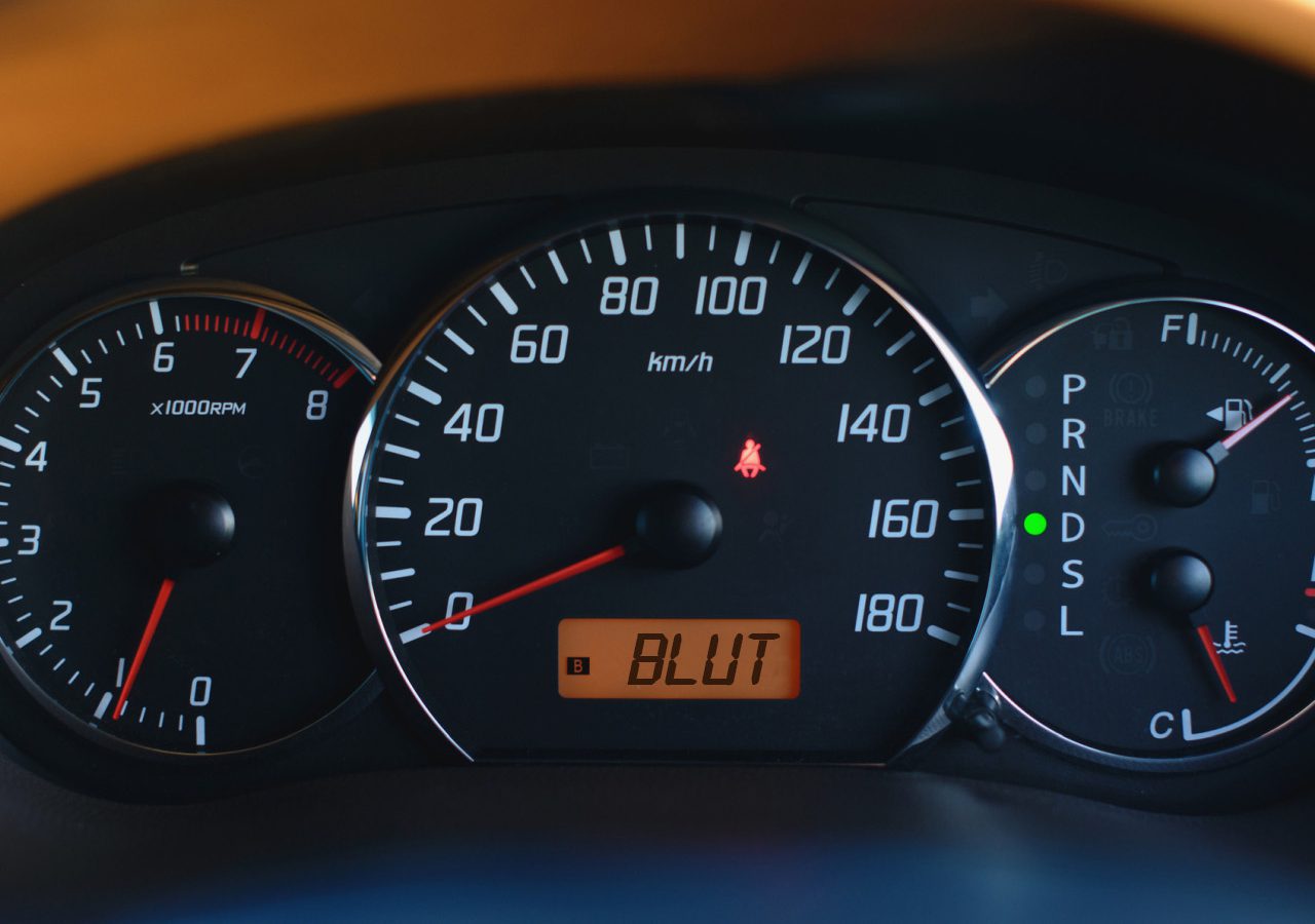 Close-up van het dashboard van een auto met de snelheidsmeter, toerenteller, brandstofmeter en versnellingsindicator. De snelheidsmeter geeft 120 kilometer per uur aan en de versnellingsindicator geeft "BLUT.