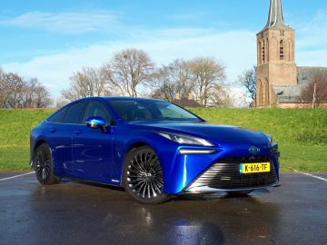 Een blauwe Toyota sedan, rijden op waterstof, staat geparkeerd op een natte stoep met groen en bomen op de achtergrond. In de verte is een kerk zichtbaar. Kentekenplaat: K-616-TF.