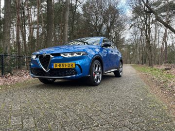 Een blauwe Alfa Romeo Tonale SUV met kenteken R-851-DR staat geparkeerd op een verhard pad in een bosrijke omgeving omgeven door bomen zonder bladeren.