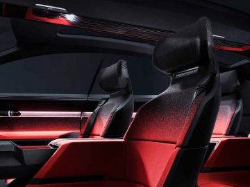 Een modern auto-interieur met twee voorstoelen met een strak ontwerp, bekleding met rode accenten en sfeerverlichting. Ook het dashboard en een gedeeltelijk zicht op de ruit zijn zichtbaar en belichamen de futuristische esthetiek van de Audi Activesphere Concept.