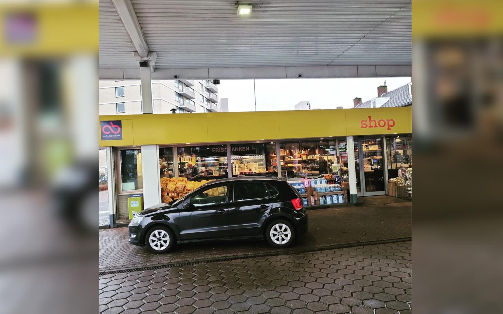 Een kleine zwarte auto staat onder een afdak geparkeerd voor de supermarkt van een tankstation. De winkel heeft grote glazen etalages waar verschillende producten tentoongesteld worden. Op het bord hierboven staat 'winkel', en verborgen in de schaduw zou je kunnen vermoeden dat er een benzinedief op jacht is.