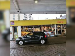 Een kleine zwarte auto staat onder een afdak geparkeerd voor de supermarkt van een tankstation. De winkel heeft grote glazen etalages waar verschillende producten tentoongesteld worden. Op het bord hierboven staat 'winkel', en verborgen in de schaduw zou je kunnen vermoeden dat er een benzinedief op jacht is.