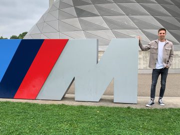Persoon die naast een groot "M"-beeld staat, dat deel uitmaakt van het BMW M-logo, buiten een futuristisch gebouw met geometrische designelementen, dat een vooruitblik biedt op autojaar 2023.