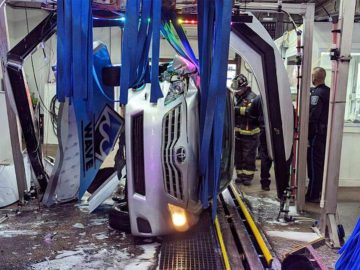 Een witte SUV verschijnt omgevallen en vastgeklemd in een wasstraat. Brandweerlieden en een politieagent zijn ter plaatse om de situatie te beoordelen te midden van felle lichten en borstels schoon te maken.