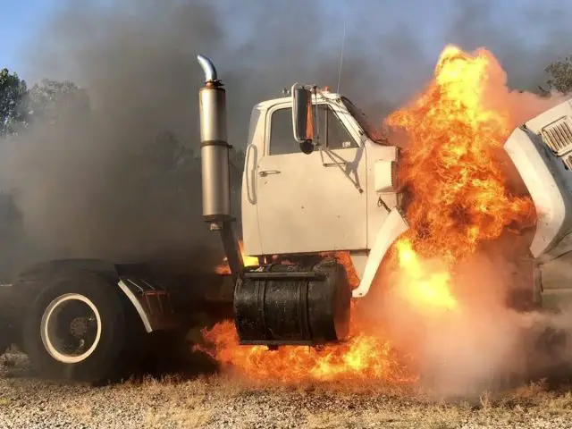 Een witte vrachtwagen met dieselmotor wordt buiten omgeven door vlammen en dikke rook, omgeven door bomen.