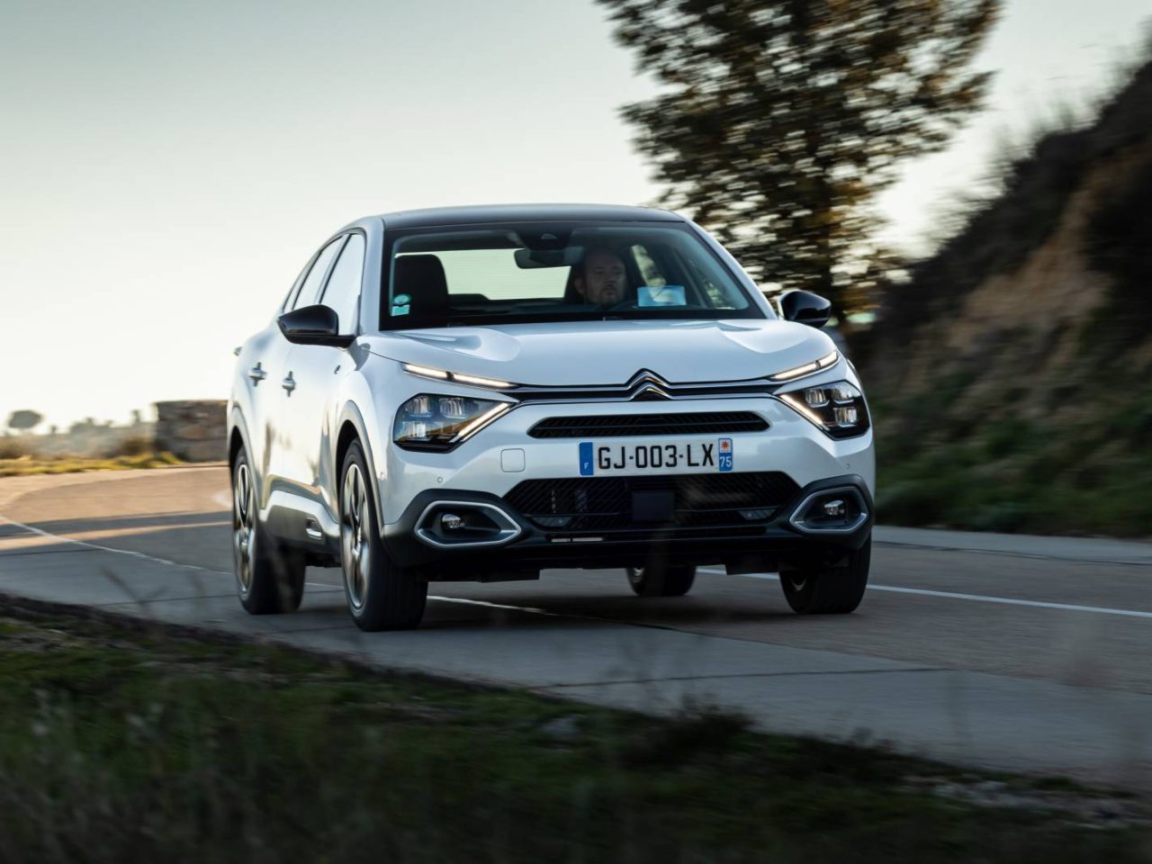 Een witte Citroën ë-C4 X glijdt over een weg, omlijst door heuvels en bomen op de achtergrond.