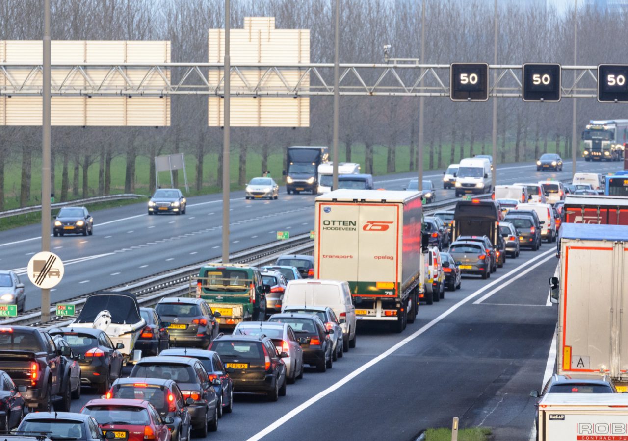 Zware verkeersopstoppingen op een snelweg met meerdere rijstroken, waar auto's en vrachtwagens overdag in de rij staan. Er strekt zich een dicht wagenpark uit, met op de achtergrond bomen en verschillende bovengrondse borden.
