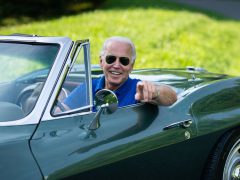 Een man in een blauw shirt glimlacht en wijst terwijl hij op de bestuurdersstoel van een vintage groene cabriolet zit, die doet denken aan de klassieke stijl die Biden bewondert. Groen gras en een weg zijn zichtbaar op de achtergrond.