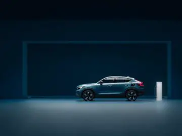 Binnen staat een blauwe elektrische Volvo SUV met achterwielaandrijving geparkeerd, aangesloten op een witte laadpaal. De achtergrond is donkerblauw en contrasteert met het goed verlichte voertuig en de oplader.