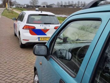 Een witte politieauto met "POLITIE" op de achterkant staat achter een blauwe auto geparkeerd in de berm, mogelijk met rijden zonder rijbewijs, met velden en gebouwen op de achtergrond.