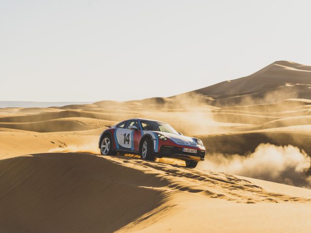 Een Porsche 911 Dakar met race-emblemen rijdt over een woestijnduin en genereert een stofspoor tegen de achtergrond van zandheuvels onder een heldere hemel.