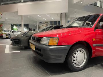 Een rode Porsche geparkeerd op de voorgrond naast een donkergrijze auto in een moderne showroom met andere voertuigen zichtbaar op de achtergrond.