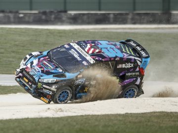 Een rallyauto bedekt met sponsorlogo's slipt op een onverharde weg en werpt stof op terwijl hij naar één kant kantelt met de voorwielen van de grond, wat een indrukwekkende Gymkhana-manoeuvre laat zien.