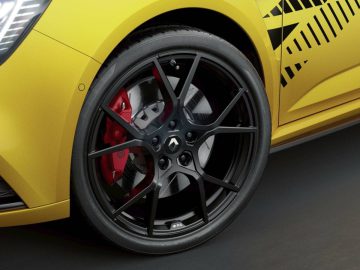 Close-up van het voorwiel van een Renault met een zwarte lichtmetalen velg en rode remklauw op een geel voertuig, met een decoratief zwart patroon boven het stuur.
