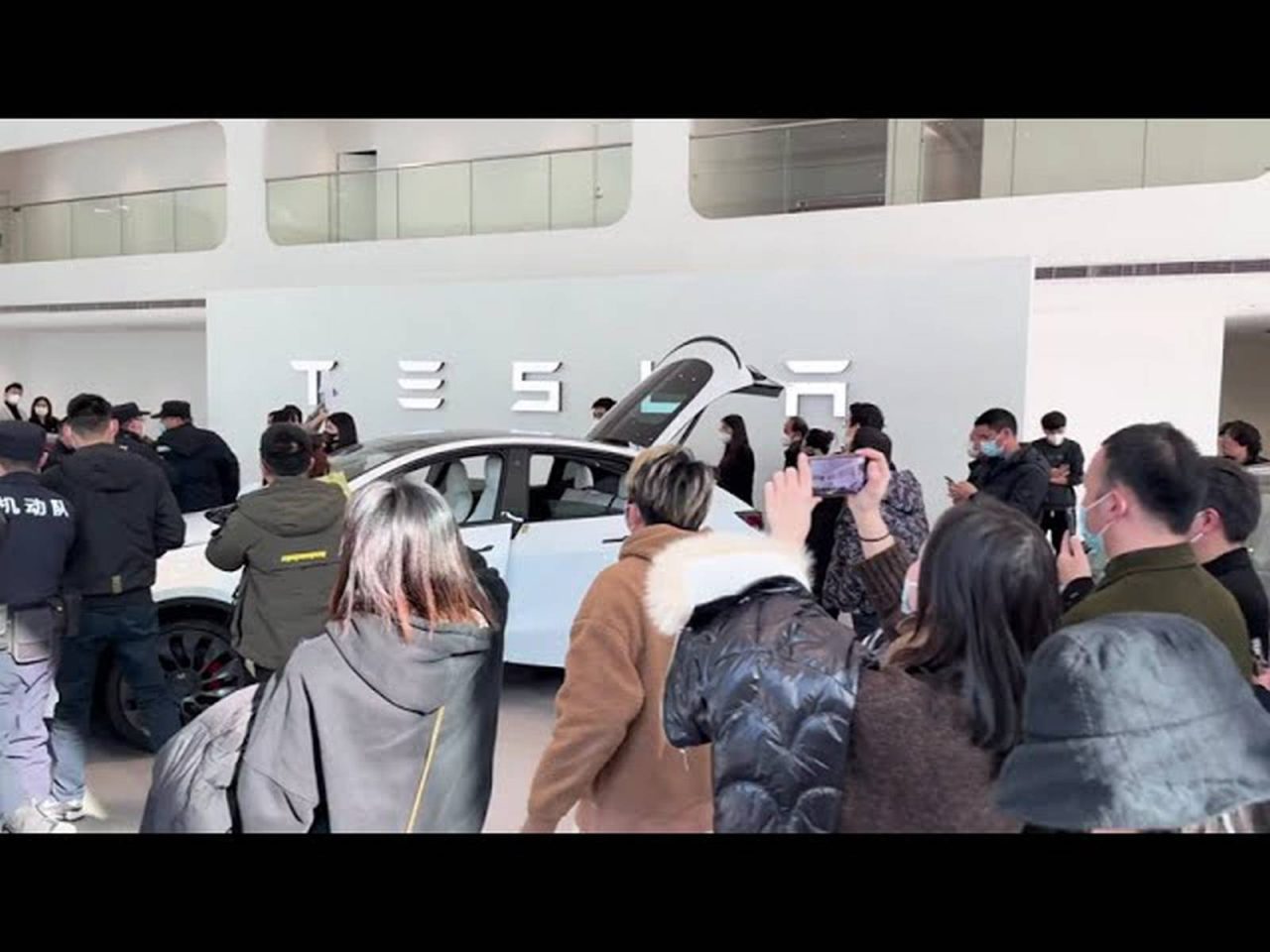 Een menigte mensen verzamelt zich rond een strakke witte Tesla in de showroom, met het iconische Tesla-logo prominent op de muur op de achtergrond.