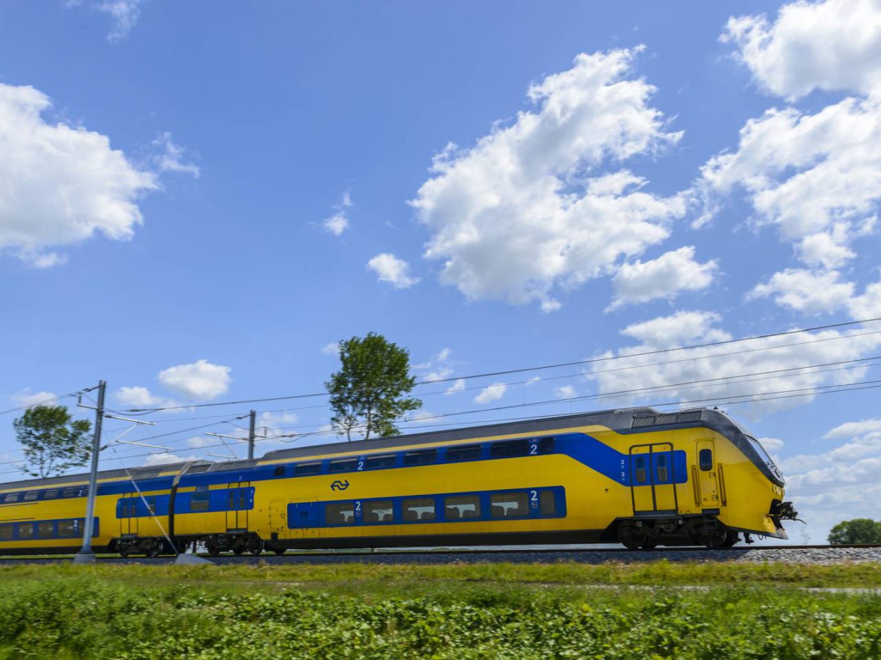 Een geel-blauwe dubbeldekker-passagierstrein, niet verzekerd, rijdt op een spoor onder een gedeeltelijk bewolkte hemel met groen gras en bomen op de achtergrond.