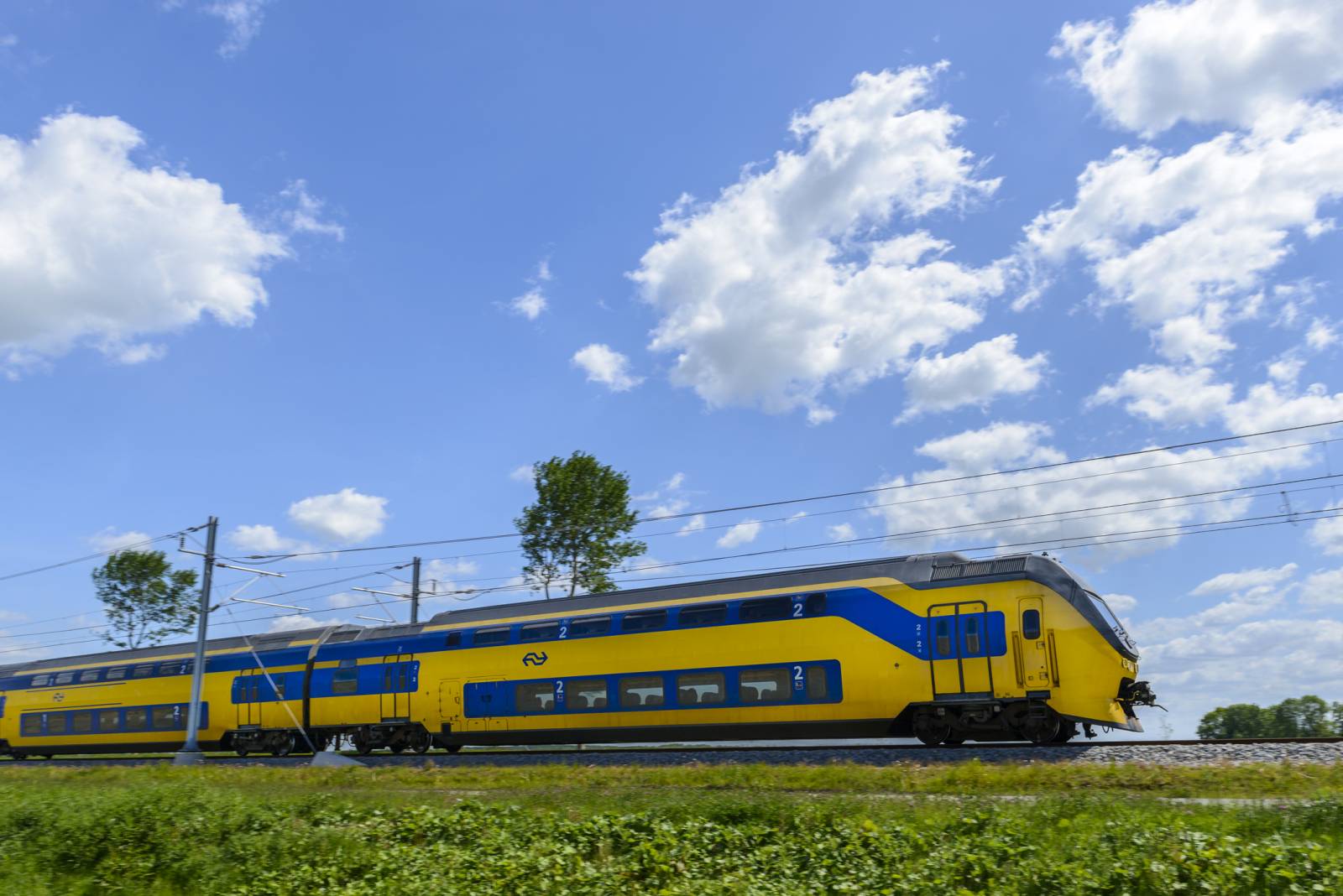 Een geel-blauwe dubbeldekker-passagierstrein, niet verzekerd, rijdt op een spoor onder een gedeeltelijk bewolkte hemel met groen gras en bomen op de achtergrond.