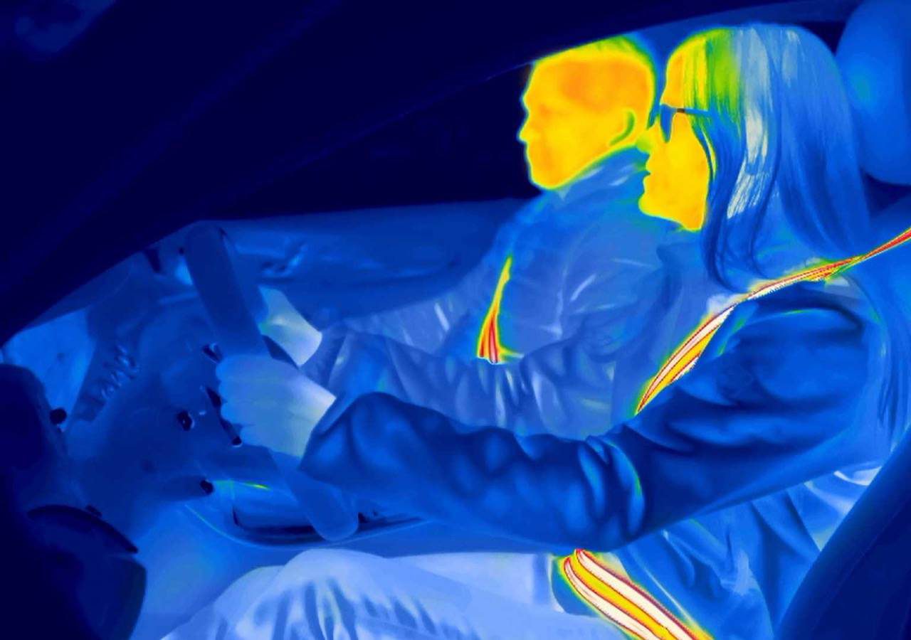 Infraroodbeeld van twee mensen in een auto. De bestuurder, met één hand aan het stuur en een bril op, is duidelijk zichtbaar. Beide personen hebben hun gordels veilig vastgemaakt. Warme kleuren duiden op hogere temperaturen.