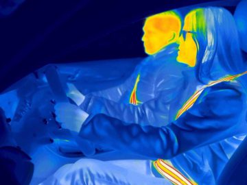 Infraroodbeeld van twee mensen in een auto. De bestuurder, met één hand aan het stuur en een bril op, is duidelijk zichtbaar. Beide personen hebben hun gordels veilig vastgemaakt. Warme kleuren duiden op hogere temperaturen.