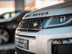 Close-up van de grille en het embleem van een witte Range Rover Evoque, met een wazig voertuig op de achtergrond. Interesse in deze luxe SUV? Vraag me af hoeveel het kost om er een te bezitten.