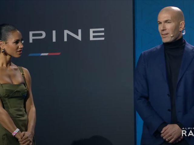 Twee personen staan voor een achtergrond met de naam en het logo van Alpine en zijn in gesprek. De persoon links draagt een olijfkleurige jurk en de persoon rechts een blauwe blazer.