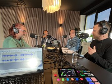 Vier mensen zitten aan een tafel versierd met podcastapparatuur, microfoons en audiomixers, bezig met een AutoRAI Podcast-opnamesessie in een goed verlichte kamer. Een laptop toont audiogolfvormen.