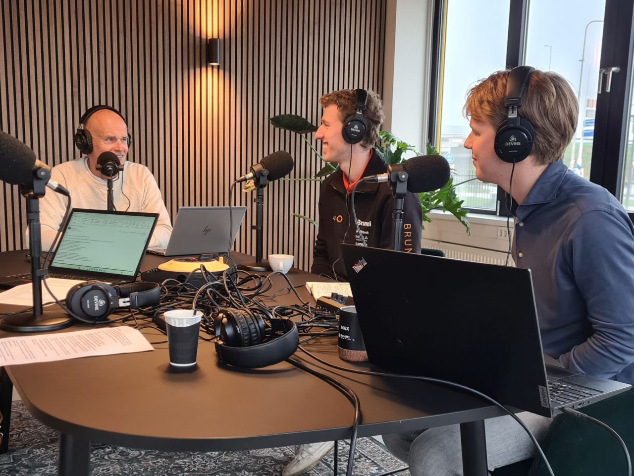 Drie personen zitten rond een tafel met microfoons, laptops en koptelefoons en zijn bezig met een AutoRAI Podcast-opnamesessie. Op de achtergrond zijn een raam en decor te zien.