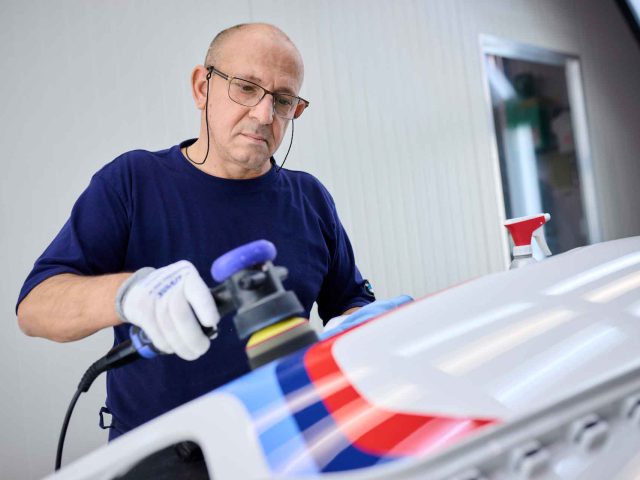 Een man met bril, handschoenen en gehoorbescherming poetst het oppervlak van een witte BMW 3.0 CSL, versierd met blauwe en rode strepen, met behulp van elektrisch polijstgereedschap in een werkplaats.