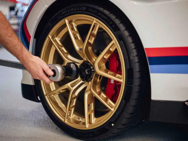 De hand van een persoon die gereedschap gebruikt om een gouden lichtmetalen velg van een hoogwaardige BMW 3.0 CSL vast te draaien. Het stuur is voorzien van Michelin Pilot Sport 4 S banden en rode remklauwen.