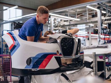 Twee arbeiders monteren een autobumper in een helder verlichte werkplaats, waarbij ze zich concentreren op het bevestigen van componenten aan het witte, rode en blauwe paneel van de BMW 3.0 CSL.