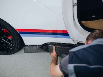 Een persoon in werkplaatsuniform is bezig met het afstellen of installeren van een zijskirt van koolstofvezel op een witte BMW 3.0 CSL met rood en blauw gestreepte details bij het achterwiel.