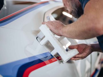 Een persoon brengt zorgvuldig een wit stencil aan op de zijkant van een BMW 3.0 CSL, die voorzien is van opvallende rode, blauwe en zwarte strepen.