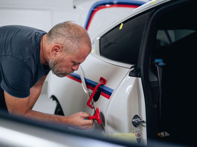 Een persoon met een baard repareert een witte BMW 3.0 CSL-carrosserie met rood zuiggereedschap.