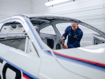 Een man in blauw uniform werkt in een helder verlichte garage nauwgezet aan het interieur van een wit BMW 3.0 CSL-frame.