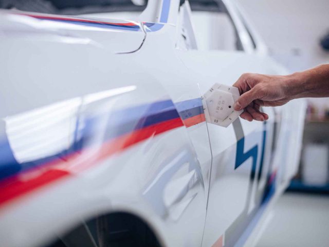 Een persoon meet de kleurnauwkeurigheid van de witte lak van een BMW 3.0 CSL door deze te vergelijken met een kleurstaal dat tegen de carrosserie van de auto wordt gehouden en waarop rode en blauwe strepen zijn aangebracht.