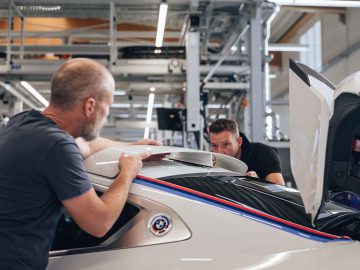 Twee mannen werken aan een witte BMW 3.0 CSL in een industriële garage, de een past de achterspoiler aan en de ander inspecteert de open achterklep van het voertuig.