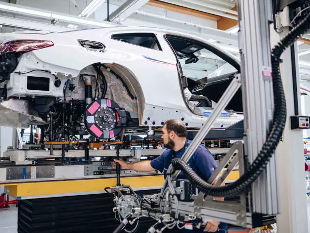 Een arbeider in een blauw shirt bedient machines terwijl hij een witte BMW 3.0 CSL monteert op een verhoogd platform in een autofabriek.