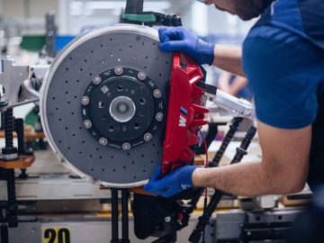 Een persoon met blauwe handschoenen monteert in een industriële omgeving minutieus een grote rode remklauw op een zilveren remschijf, die doet denken aan de precisie die vereist is voor een BMW 3.0 CSL.