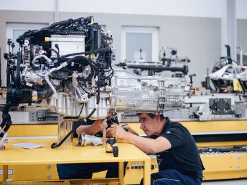 Een technicus werkt in een werkplaats aan een groot motoronderdeel, omringd door machines en gereedschappen, en monteert de onderdelen net zo nauwgezet als bij de iconische BMW 3.0 CSL.