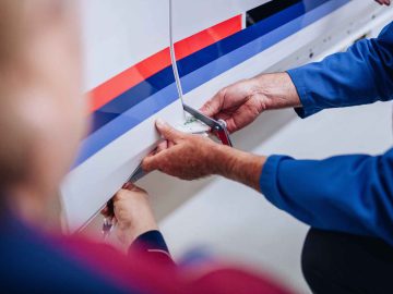 Twee mensen in blauwe shirts werken samen om een bout vast te draaien op een wit oppervlak met rode en blauwe strepen, dat doet denken aan de klassieke BMW 3.0 CSL racekleuren.