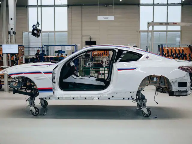 Een gedeeltelijk geassembleerde witte BMW 3.0 CSL-carrosserie op een productielijn in een fabriek, waarbij deuren en verschillende onderdelen ontbreken, geplaatst op een set standaards voor stabiliteit.