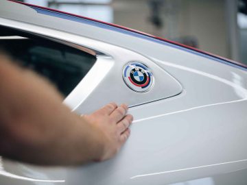 De hand van een persoon raakt de zijkant van een witte auto aan, waarbij de nadruk ligt op het BMW-logo bij de achterruit, dat doet denken aan de iconische BMW 3.0 CSL.