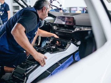 Een persoon in een werkplaats is ijverig bezig met het installeren of repareren van de middenconsole van het interieur van een BMW 3.0 CSL. Gekleed in een blauw uniform gebruiken ze vakkundig gereedschap op het dashboard, zodat elk detail aan hoge normen voldoet.