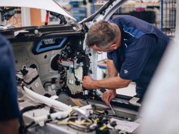 Een monteur in blauw uniform repareert minutieus de interne onderdelen van een gedeeltelijk gedemonteerde BMW 3.0 CSL.