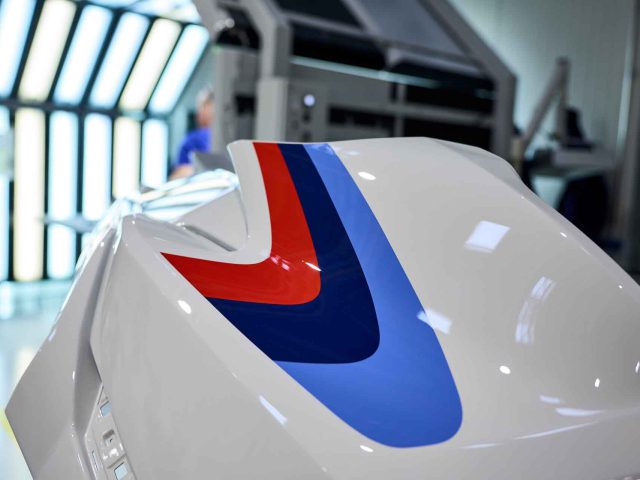 Close-up van het gebogen oppervlak van een witte BMW 3.0 CSL met rode en blauwe strepen in een helder verlichte autowerkplaats.