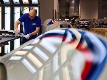 Een arbeider in een fabriek, gekleed in een blauw shirt en witte handschoenen, inspecteert een vliegtuigonderdeel onder fel licht, net zoals de nauwgezette precisie die vereist is bij het restaureren van een klassieke BMW 3.0 CSL.