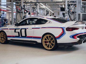 Een BMW 3.0 CSL, wit met blauwe, rode en zwarte strepen, het nummer 50 op de zijkant en gouden wielen staat geparkeerd in een goed verlichte garage.