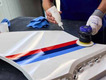 Persoon die een polijstmachine gebruikt op een wit BMW 3.0 CSL-onderdeel met rode, blauwe en lichtblauwe strepen terwijl hij polijstmiddel aanbrengt.