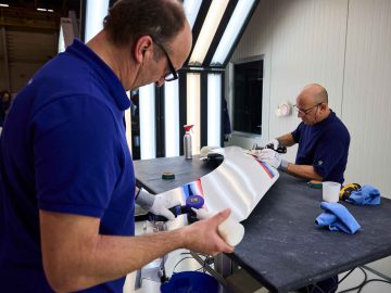 Twee mannen in blauwe overhemden poetsen en werken minutieus aan een glimmende BMW 3.0 CSL in een goed verlichte werkplaats. De ene man concentreert zich met gereedschap op de auto, terwijl de ander materialen klaarmaakt.